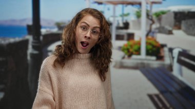 Gözlüklü bir kadın, deniz kenarında güneşli bir günde etrafı manzarayla çevrili, rahat bir kazak giymiş, kumsal arkaplanı ve canlı çiçekleri olan bir kadın..