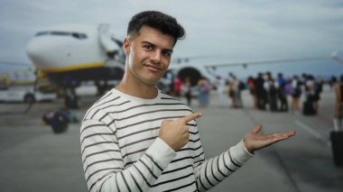 Havaalanındaki Latin asıllı genç adam hava alanının arka planında rahatlamış seyahat anını gösteren jestler yapıyor.