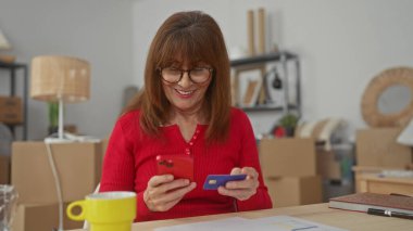 Kutularla çevrili yeni apartman dairesinde akıllı telefon ve kredi kartı kullanan İspanyol bir kadın taşınma ya da yenileme önerisi sunuyor..