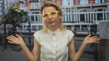 Sarı gözlüklü kadın, şehir binası cephesi altında sıradan bir omuz silkme hareketiyle sokakta el kaldırıyor; belirsizlik.