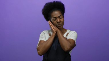 Önlük giyen Afrikalı Amerikalı kadın mor arka planda uyuma hareketi yapıyor. Bu da huzurlu bir ortamda yorgunluk olduğunu gösteriyor..