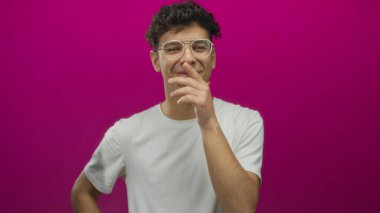 Gözlüklü genç adam kameraya ve ağıza bakan beyaz tişörtlü eflatun stüdyoda gülüyor..