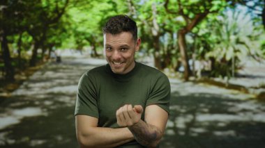 Genç adam açık hava parkında gülümsüyor ağaçlar ve güneş ışığı dallardan süzülüyor, dövmeli kolunu ve günlük tarzını gösteriyor..