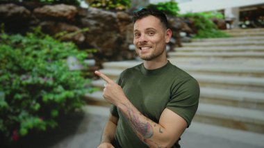 Açık havada gülümseyen bir adam yeşil bahçeyi işaret ediyor. Mutluluk ve günlük giysiler sergiliyor. Dövmeleri ve kısa saçı olan bir şehir zeminine karşı..