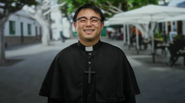 Gözlüklü ve haçlı siyah cüppeli gülümseyen rahip şehir caddesinde ağaçların ve açık hava kafelerin bulanık arka planıyla duruyor..