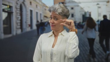 Beyaz kadın, güneşli bir kaldırım taşı caddesinde kol saatinin yanında parmak uçlarını sıkıyor; şüphecilik.