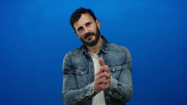 Sakallı, kot tişörtlü ve beyaz tişörtlü İspanyol bir adam mavi arka planda belirgin jestler yapıyor..