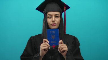 Başlık ve cüppeli mezun bir kadın, izole edilmiş yeşil arka plana karşı Ukrayna pasaportu taşıyor. Bu da yeni akademik seyahat ve seyahat fırsatlarını simgeliyor..