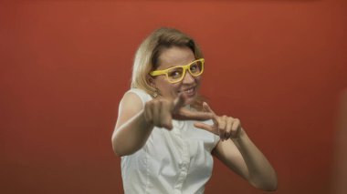 Sarı gözlüklü ve beyaz kolsuz bir kadın stüdyoda kameraya parmağıyla işaret ediyor..