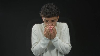 Siyah bir stüdyoda kollarını göğsünde kavuştururken gözlüklü ve beyaz kazaklı bir adam titriyor..