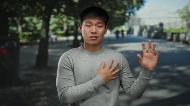 Gri kazaklı genç Çinli adam, yeşil ağaçlar ve gölgelerle çevrili güneşli bir parkta el hareketleriyle duygularını ifade ediyor..