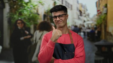 Gözlüklü genç adam, şehir caddesinde hareketli bir baş parmağıyla hareketli hareketler yapıyor. Etrafı bulanık gezginler ve şehir yeşillikleriyle çevrili..