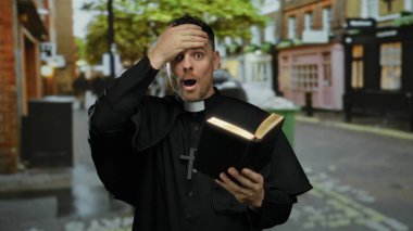 Şehir sokaklarında kitap okuyan rahip, şok olmuş bir ifadeyle siyah bornoz giyiyor ve şehrin bulanık arka planında ağaçlar ve binaları gösteren çapraz bir kolye takıyor.