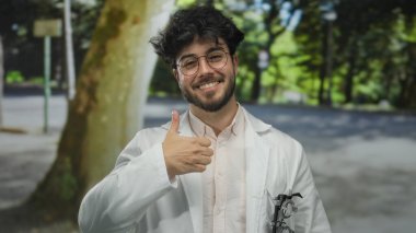 Sakallı, gözlüklü ve beyaz önlüklü İspanyol bir adam yeşil bir açık hava parkında baş parmağını kaldırıyor..