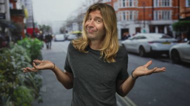 Uzun sarı saçlı bir adam kırmızı tuğla binalarla kaplı kalabalık bir şehir sokağında omuzlarını silker; belirsizlik.