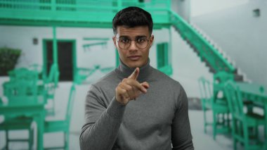 Dışarıda terasta jestler yapan genç bir adam, gözlüklü ve gri kazaklı, yeşil sandalyeli ve masalı bir şehir restoranının arka planında.