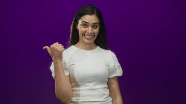 Mor stüdyoda gülümseyen ve başparmağıyla işaret eden İspanyol genç kadın; güven iyimserliği öneri yönü.