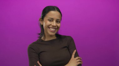 Canlı pembe bir arka planda, kollarını kavuşturarak güvenle gülümseyen bir kadın, stüdyo ortamında mutluluk ve pozitifliği somutlaştıran, günlük kahverengi bir gömlek giyen bir kadın..