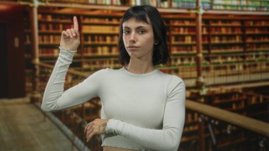 Elleri açıkta olan kadın, kütüphane binasındaki ahşap kitaplığa doğru işaret ediyor; merak uyandıran keşif keşfi.