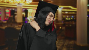 Mezuniyet elbisesi giymiş genç bir kadın kumarhanede renkli kumar makineleriyle omuz ağrısı çekiyor..