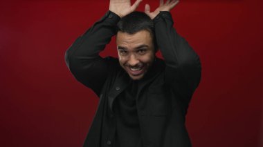 Kırmızı bir stüdyoda siyah tişörtlü bir adam sırıtıyor ve ellerini başının üstüne kaldırıyor..