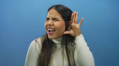 Uzun saçlı bir kadın, izole edilmiş mavi bir arka plana dikkatlice kulak veriyor. Beyaz bir kazak giyiyor ve merakını ve dikkatini gösteriyor..