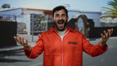 Turuncu hapishane tulumlu İspanyol bir adam sokakta dikiliyor, bir binanın arka planına karşı şaşkınlık ve duygularını ifade ediyor..