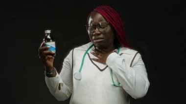 Kırmızı örgülü ve steteskoplu Afro-Amerikan kadın bir şurup şişesi tutuyor ve boğazına dokunuyor, siyah bir arka planda izole edilmiş, boğaz ağrısı için ilaç öneriyor..