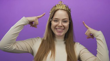 Taçlı ve gözlüklü genç bir kadın gülümsüyor ve mor stüdyoda parmağıyla tacı işaret ediyor..
