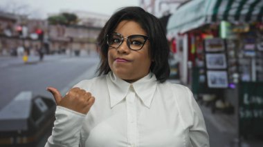 Meşgul şehir caddesinde, gözlüklü ve beyaz tişörtlü bir kadın, şehir aktivitelerinin arka planı ve hareketli açık hava ortamlarıyla belirsizlik gösteriyor..