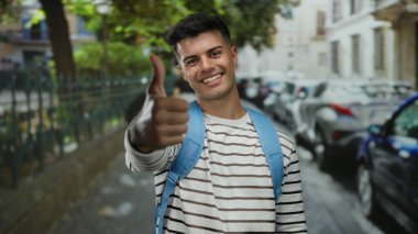 Şehir caddesinde başparmağıyla gülümseyen genç bir adam, çizgili bir gömlek ve mavi bir sırt çantası, park etmiş arabalar ve ağaçlarla çerçevelenmiş, dışarıda pozitiflik yayıyor..