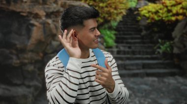 Genç adam dışarıda kulağına gülümsüyor ve el kol hareketi yapıyor. Arka planda merdivenleri olan çizgili gömlek ve sırt çantası giyiyor..