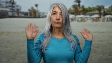 Mavi kazaklı, gri saçlı yaşlı kadın güneşli bir deniz kenarında duruyor. Palmiye ağaçları ve şemsiyelerin yer aldığı bir kumsal zeminine karşı ellerini kaldırarak ifade ediyor..