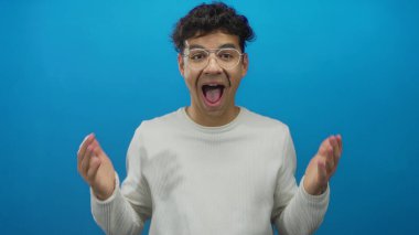 Beyaz eşofman altı giyen İspanyol genç adam ağzını açar ve mavi stüdyoda elleri yanaklarına bastırır..