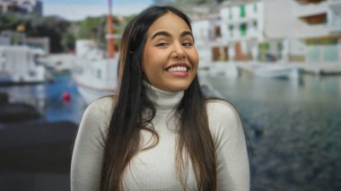 Beyaz kazaklı, gülümseyen İspanyol kadın arka planda tekneler ve binalar olan bir deniz kenarında..