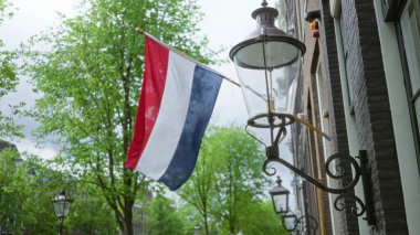 Hollanda bayrağı, yemyeşil ağaçlar ve bulutlu bir günde geleneksel sokak lambalarıyla çevrili Amsterdam 'daki bir binanın önünde asılı duruyor..