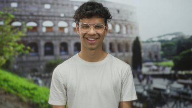 Beyaz tişört giyen ve gözlüklü genç İspanyol adam Roma binasında görünen yüzüyle gülümsüyor. Mutluluk..
