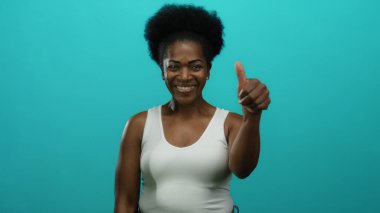 İzole edilmiş mavi arka planda güvenle gülümseyen bir kadın olumlu ve mutluluk saçan bir başparmak verir..