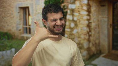 Adam taş bina girişinin önünde serçe parmağı ve baş parmağıyla telefon hareketi yapıyor; dostça iletişim.