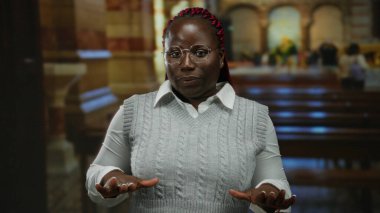 Kilise ortamında gözlüklü ve gri kazaklı Afro-Amerikalı bir kadın, sakin görünüyor, elleri açık, geleneksel kilise sıraları ve hafif aydınlatma ile çevrili..