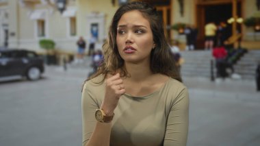 Şehir sokak ortamında endişeli bir ifadeyle dışarıda bir kadın, saat takıyor, arka planda insanlar hareketli bir şehir atmosferini işaret ediyor..