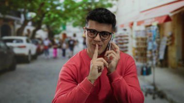 Genç adam dışarıda akıllı telefonda konuşurken gülümsüyor, canlı bir şehir caddesinde ağaçlarla dikiliyor ve arka planda insanları bulanıklaştırıyor, gözlük takıyor ve kırmızı bir kazak giyiyor..