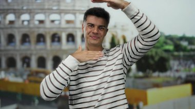 Roma ikonik stadyumunun önünde neşeli bir şekilde poz veren genç adam, açık bir gülümseme ve çizgili bir gömlek sergiliyor, canlı bir şehir atmosferini yakalıyor..