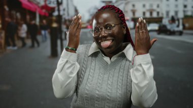 Şehirli bir kadın şakacı bir şekilde elleri ile yüzünü örtüyor sonra da canlı şehir arkaplanıyla geniş bir şekilde gülümsüyor..