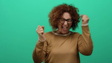 Kıvırcık saçları ve gözlükleriyle duygularını gösteren, kahverengi bir gömlek giyen ve çerçevede izole edilmiş bir kadın, canlı yeşil bir arka plana karşı ifade edici bir poziyonda parmaklarıyla jestler yapıyor..