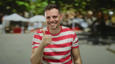 Açık havada gülümseyen genç adam şehir sokaklarında arka planda bulanık, çizgili tişört giyen, şehir ortamında kendine güven ve pozitiflik gösteren baş parmağını kaldırıyor..