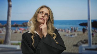 Düşünceli kadın yargıç üniforması giymiş, açık havada güneşli bir günde okyanus manzaralı bir plajda durup derin düşüncelere dalmış.