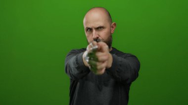 Kel, beyaz, sakallı, yeşil arka planda parmak silahlarıyla poz veren kararlı ve odaklanmış bir adam.