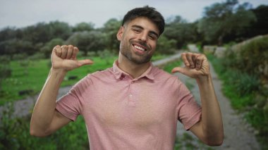 Yeşil tarlalar tarafından çerçevelenmiş bir orman yolu boyunca pembe bir polo tişörtle adam gülümser ve her iki başparmak kendine işaret eder; gurur.