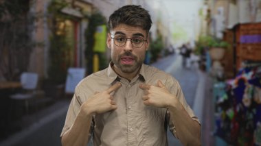 Gözlüklü, gülümseyen sakallı adam, güneşli bir şehir sokağında yumruklarını sıkar, dükkanlar ve gezgin bulanıklığıyla; sevinç zaferi.